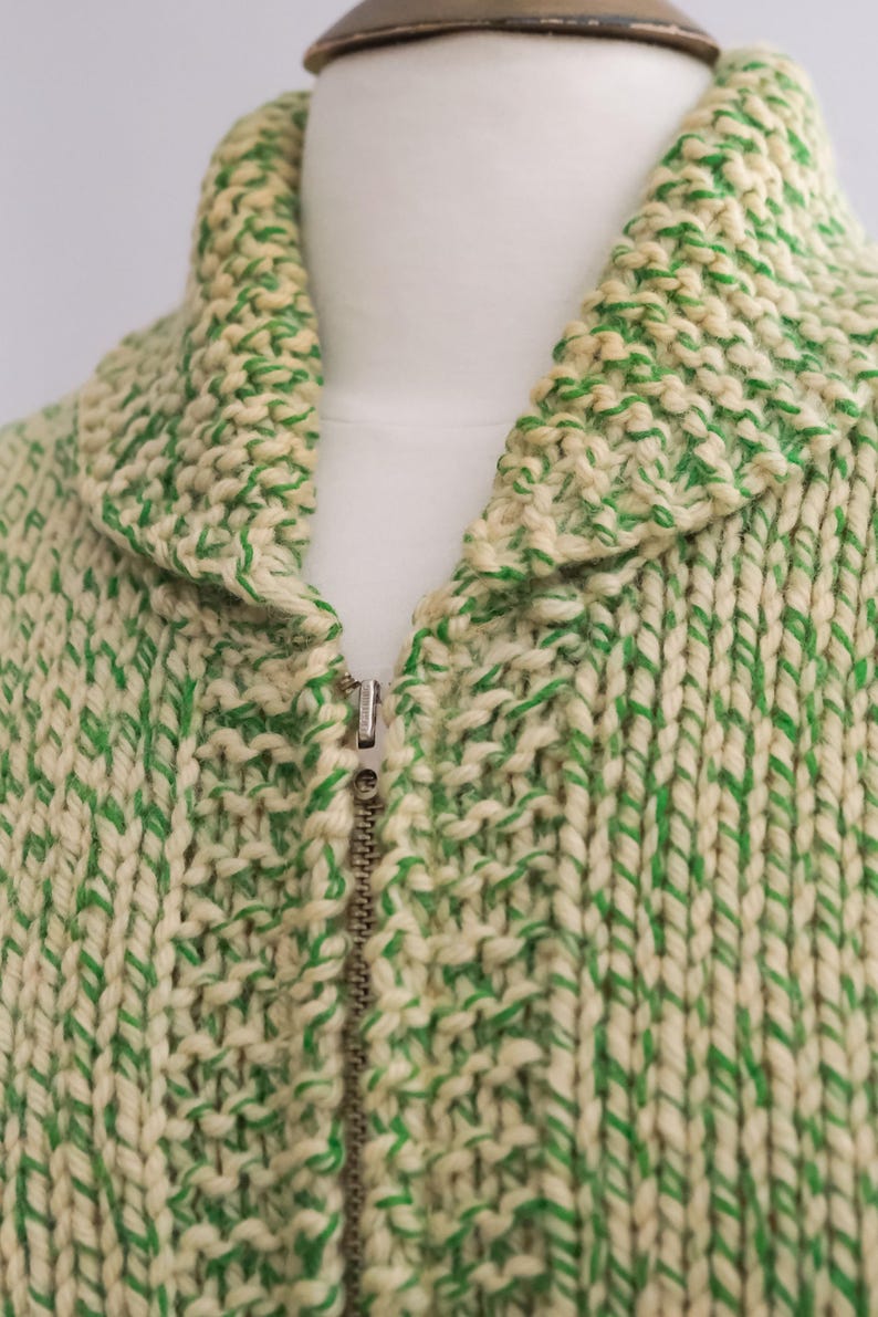 May include: Close-up of a cream-colored knitted jacket with green accents. The jacket features a high collar and a full zipper closure. The thick knit creates a textured appearance, and the green yarn is interspersed throughout the cream base.