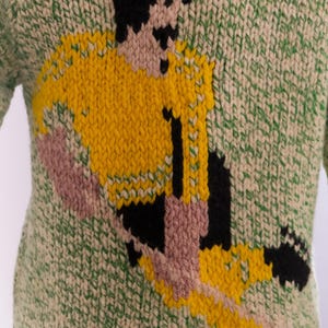 May include: Green and cream-colored knitted sweater featuring a soccer player in yellow, black, and pink. The soccer player is in action, with the number 11 on the jersey. The sweater has a textured knit pattern.