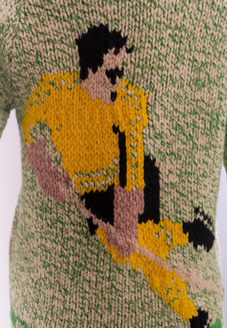 May include: Green and cream-colored knitted sweater featuring a soccer player in yellow, black, and pink. The soccer player is in action, with the number 11 on the jersey. The sweater has a textured knit pattern.