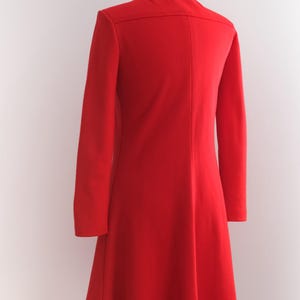 Op de afbeelding: Een felrode jas met lange mouwen en een uitlopende rok, gepresenteerd op een mannequin. De jas heeft een klassieke kraag en een getailleerd silhouet, wat een vintage stijl laat zien. De heldere kleur maakt het een opvallend kledingstuk.
