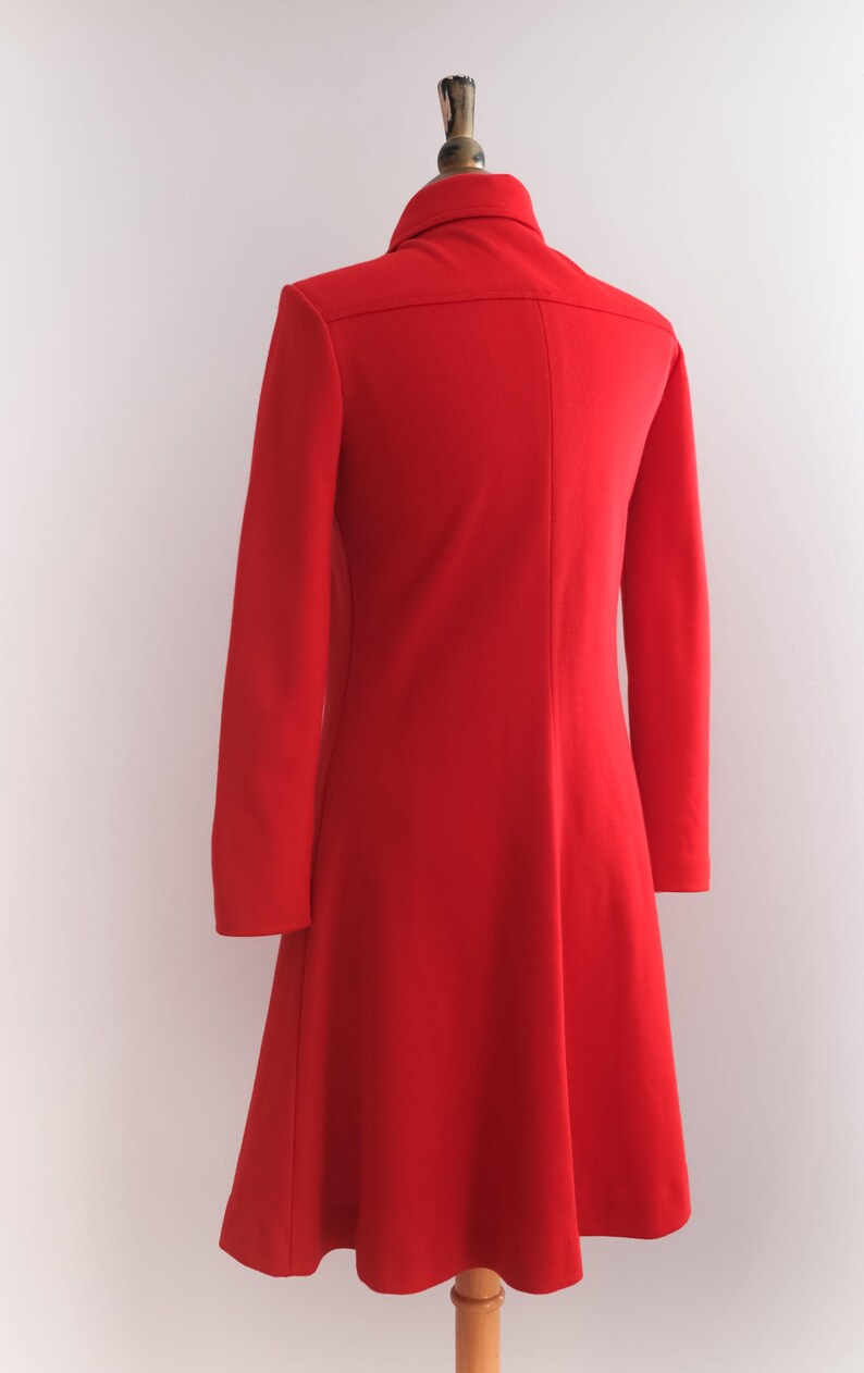 Op de afbeelding: Een felrode jas met lange mouwen en een uitlopende rok, gepresenteerd op een mannequin. De jas heeft een klassieke kraag en een getailleerd silhouet, wat een vintage stijl laat zien. De heldere kleur maakt het een opvallend kledingstuk.