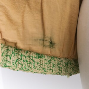 May include: Close-up of a tan fabric with a green and cream-colored knitted trim. The fabric has a slightly textured appearance and some dark staining. The knitted trim is a thick, textured band.