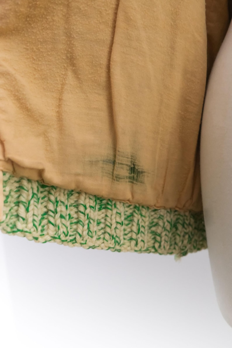 May include: Close-up of a tan fabric with a green and cream-colored knitted trim. The fabric has a slightly textured appearance and some dark staining. The knitted trim is a thick, textured band.