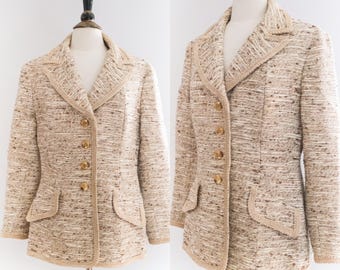 May include: A beige tweed blazer with gold buttons and a contrasting brown trim. The blazer has a double-breasted closure and flap pockets.