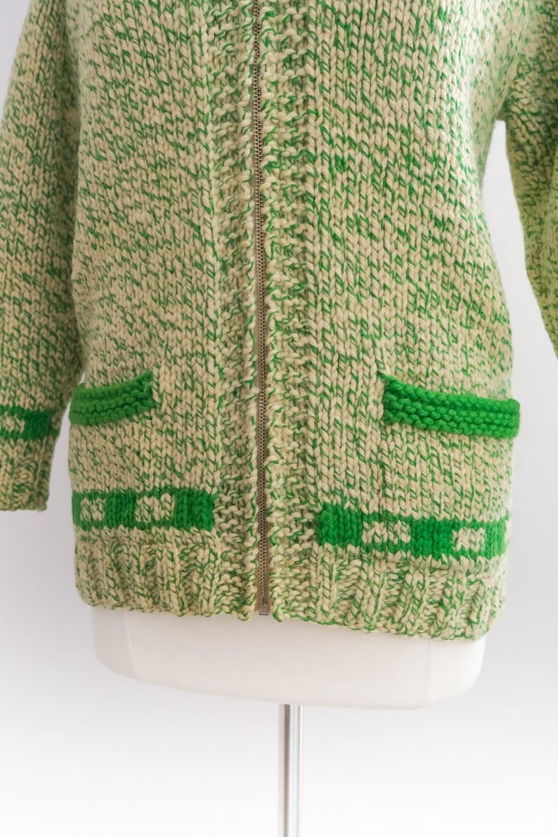 May include: A vintage green and cream-colored knitted cardigan with a full zipper closure. The sweater features two front pockets and green trim details. The knitwear has a chunky texture.