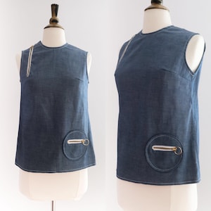 May include: A sleeveless denim top in a medium blue shade. It has a round neckline and a vertical zip detail on the left. A circular pocket with a white zip and gold-tone hardware is on the lower front. The top is displayed on a mannequin.
