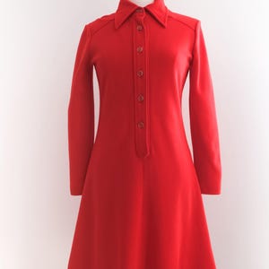 Op de afbeelding: Een felrode jurk met lange mouwen en een kraag. De jurk heeft een knoopsluiting aan de voorkant en een uitlopende rok, gepresenteerd op een mannequin. Het vintage design is een klassiek, tijdloos stuk.