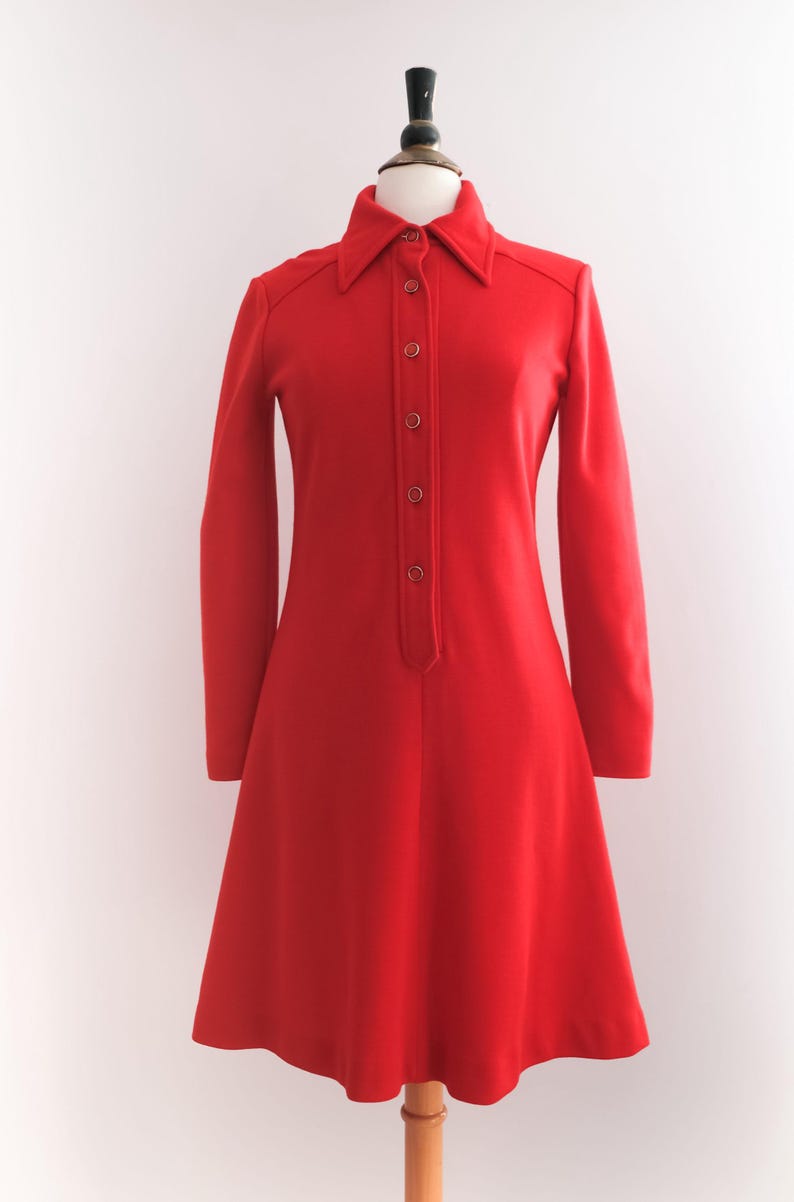 Op de afbeelding: Een felrode jurk met lange mouwen en een kraag. De jurk heeft een knoopsluiting aan de voorkant en een uitlopende rok, gepresenteerd op een mannequin. Het vintage design is een klassiek, tijdloos stuk.