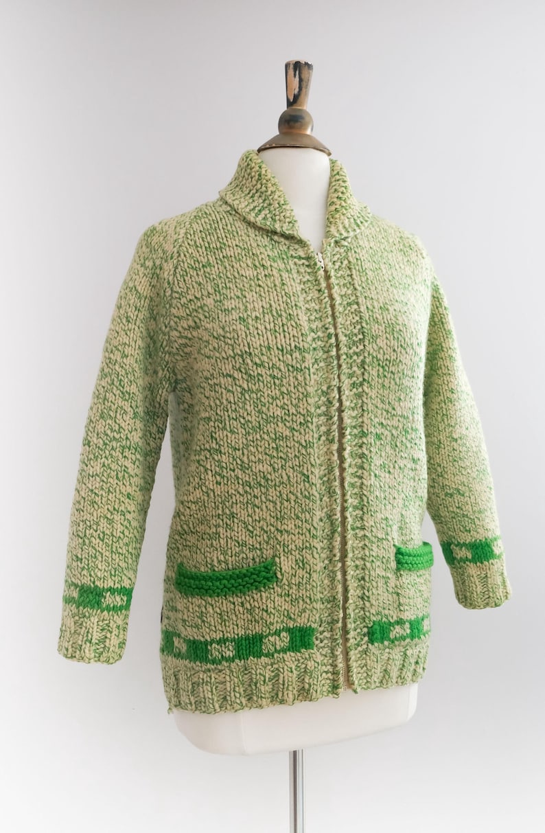 May include: A vintage green and cream-colored knitted cardigan with a full zipper closure. The sweater features two front pockets and green trim on the cuffs and bottom hem. The cardigan is displayed on a mannequin.