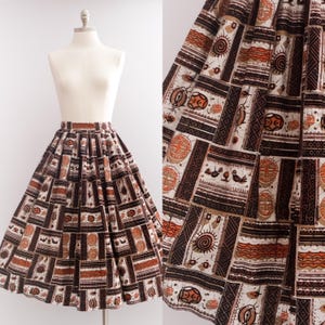 May include: A full-length skirt featuring a repeating geometric and figurative pattern in shades of brown, orange, and cream. The design includes circular motifs, stylized birds, and linear borders arranged in a structured, panel-like layout.
