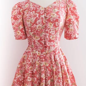 May include: A vintage-style dress with a floral pattern in shades of red, pink, and white. The dress features a V-neckline, short puffed sleeves, and a button-down front. The skirt is pleated, creating a flowing silhouette.