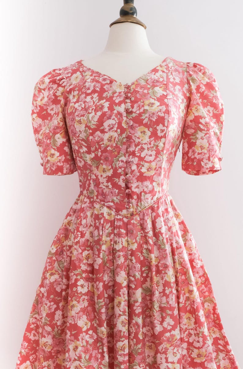 May include: A vintage-style dress with a floral pattern in shades of red, pink, and white. The dress features a V-neckline, short puffed sleeves, and a button-down front. The skirt is pleated, creating a flowing silhouette.