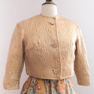May include: A light tan, cropped jacket with a textured, wave-like pattern. The jacket has three round buttons and three-quarter sleeves. The jacket is displayed on a mannequin and paired with a floral print skirt.