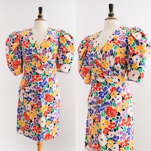 Può includere: Un abito vintage avvolgente con stampa floreale colorata. L'abito ha maniche a sbuffo e una gonna aderente. L'abito è bianco con fiori rossi, gialli, blu, verdi e viola.