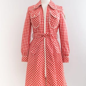 May include: A red and white gingham dress with a white zipper down the front and a white belt. The dress has two large pockets on the front and a small patch on the left side.