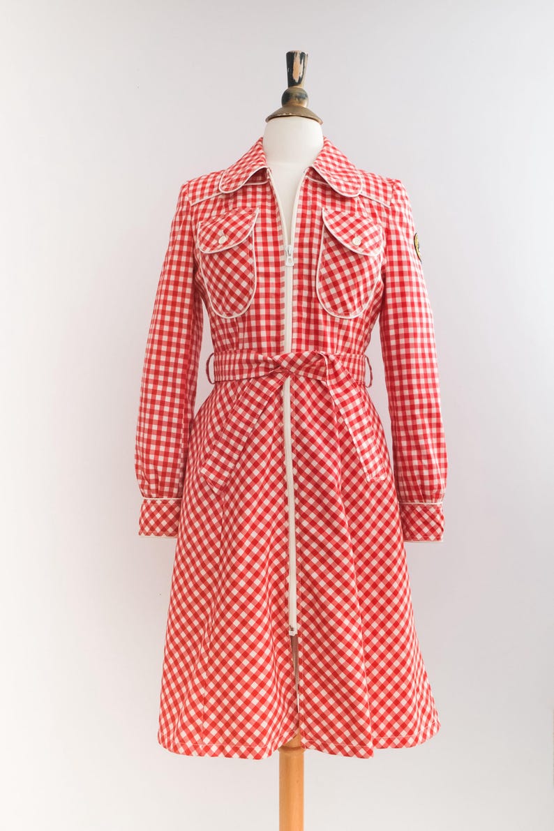 May include: A red and white gingham dress with a white zipper down the front and a white belt. The dress has two large pockets on the front and a small patch on the left side.