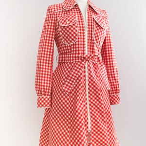 May include: A red and white gingham dress with a white zipper down the front and a white belt. The dress has long sleeves and a collar.