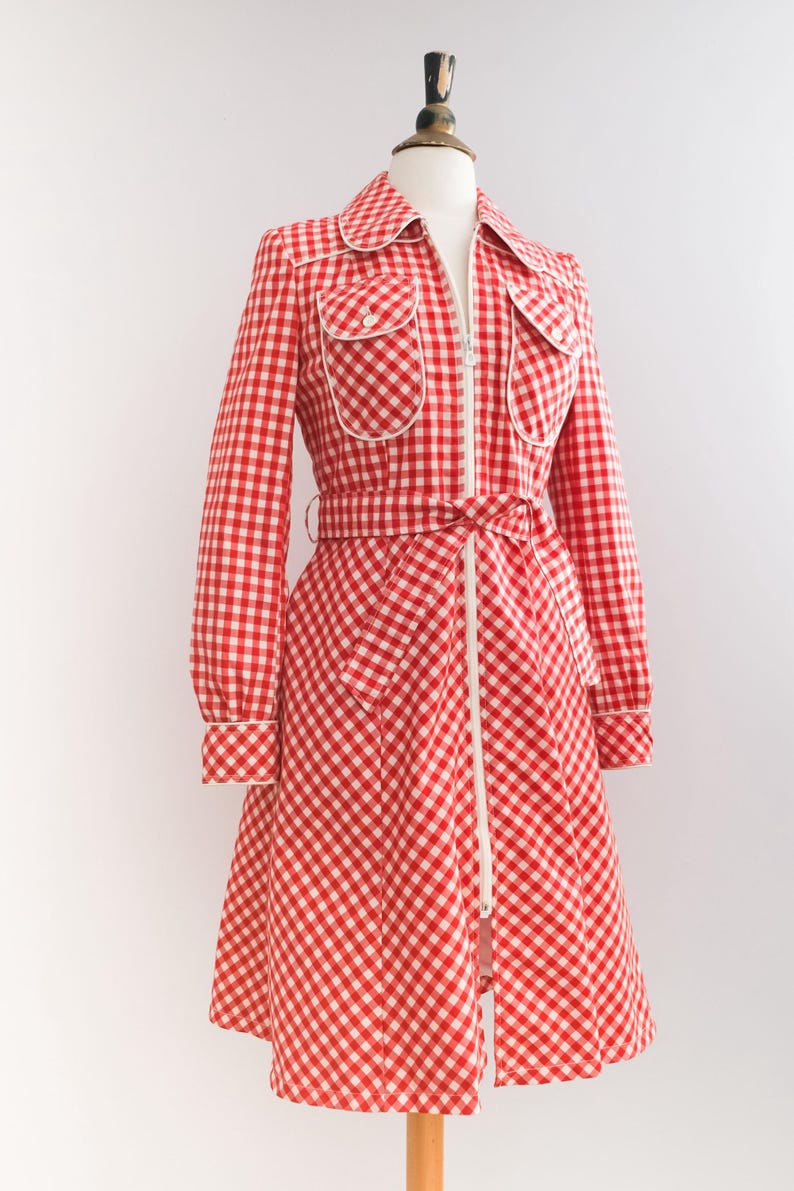 May include: A red and white gingham dress with a white zipper down the front and a white belt. The dress has long sleeves and a collar.