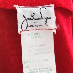 Op de afbeelding: Close-up van een rood kledingstuk met een wit label. Het label luidt "Jonathan Logan by Mel Ross Ltd." en bevat details over de stofsamenstelling en maat 13/14. Het kledingstuk is gemaakt van 50% wol en 50% acryl.