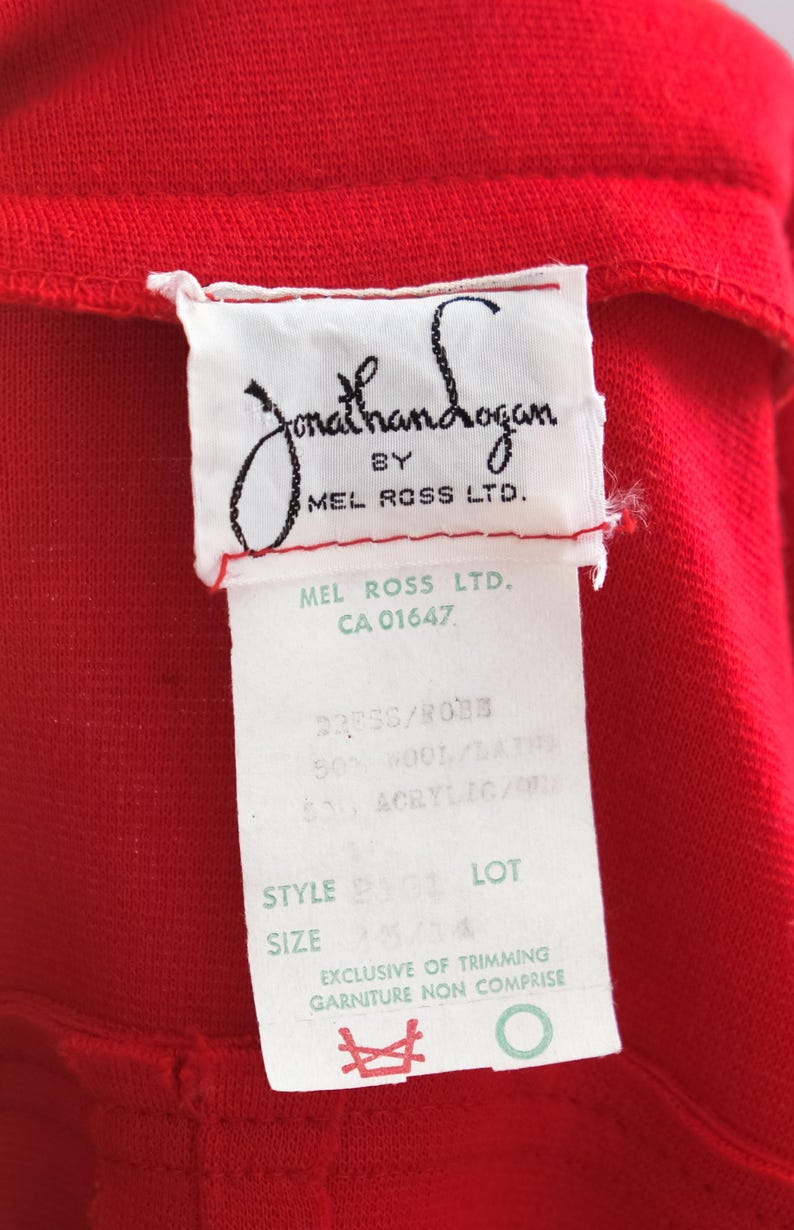 Op de afbeelding: Close-up van een rood kledingstuk met een wit label. Het label luidt "Jonathan Logan by Mel Ross Ltd." en bevat details over de stofsamenstelling en maat 13/14. Het kledingstuk is gemaakt van 50% wol en 50% acryl.