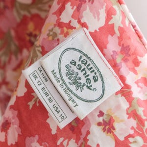 May include: Close-up of a floral fabric with a red and white pattern. The fabric has a tag that reads "laura ashley" and "Made in Hungary". The tag also includes size information: UK 12, EUR 38, USA 10.