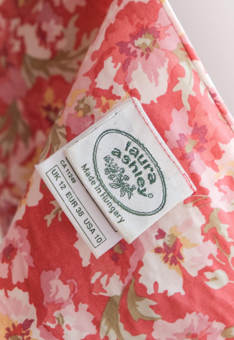 May include: Close-up of a floral fabric with a red and white pattern. The fabric has a tag that reads "laura ashley" and "Made in Hungary". The tag also includes size information: UK 12, EUR 38, USA 10.