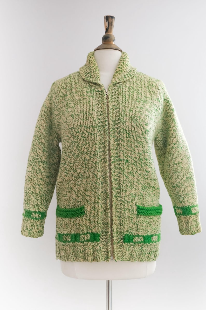 May include: A vintage green and cream-colored knit cardigan with a zipper closure. The sweater features two front pockets and green detailing on the cuffs and pockets. The collar is a shawl style.