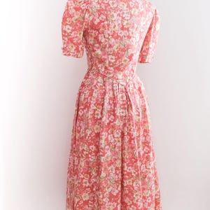 May include: A vintage-style floral dress in shades of coral, pink, and white, displayed on a mannequin. The dress features short sleeves, a fitted bodice, and a flowing skirt. The fabric is covered in a delicate floral pattern.