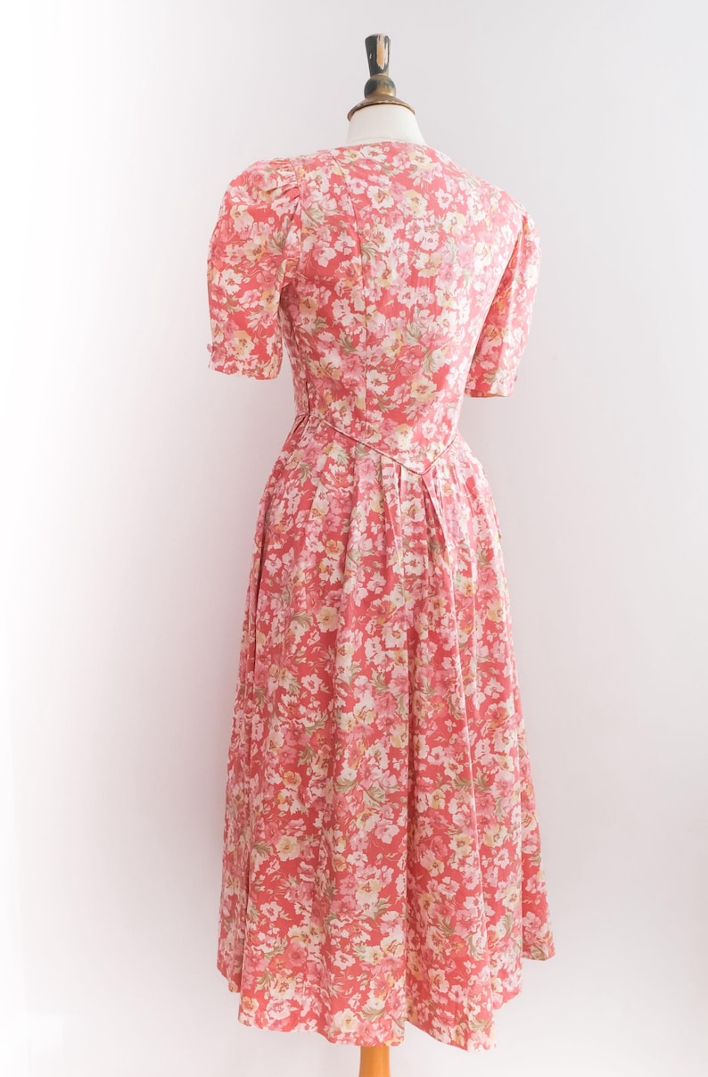 May include: A vintage-style floral dress in shades of coral, pink, and white, displayed on a mannequin. The dress features short sleeves, a fitted bodice, and a flowing skirt. The fabric is covered in a delicate floral pattern.