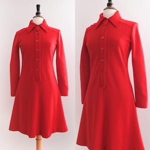 Op de afbeelding: Een felrode vintage jurk met lange mouwen en een kraag. De jurk heeft een knoopsluiting aan de voorkant en een uitlopende rok, gepresenteerd op een mannequin. De jurk is effen van kleur en toont een klassieke retro-stijl.