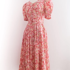 May include: A vintage-style dress with a floral pattern. The dress features a V-neck, short puffed sleeves, and a fitted bodice that flows into a pleated skirt. The fabric is a coral color with a delicate pattern of white and yellow flowers.