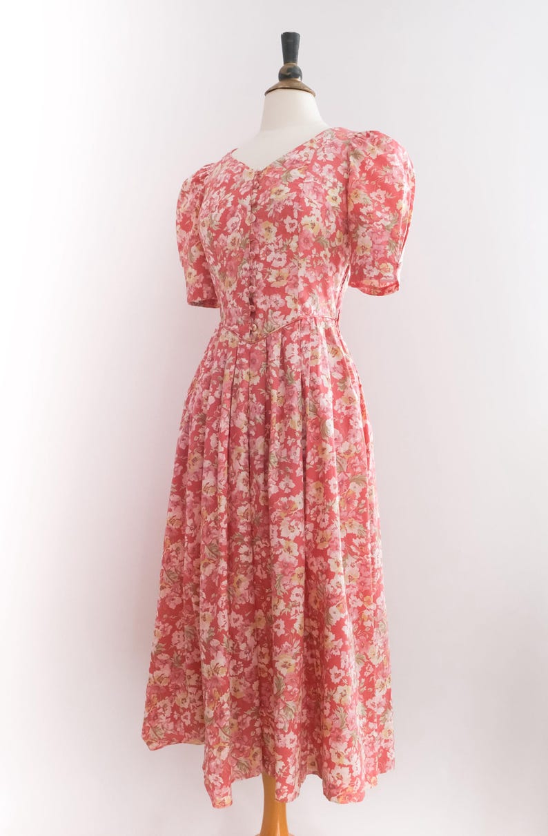 May include: A vintage-style dress with a floral pattern. The dress features a V-neck, short puffed sleeves, and a fitted bodice that flows into a pleated skirt. The fabric is a coral color with a delicate pattern of white and yellow flowers.