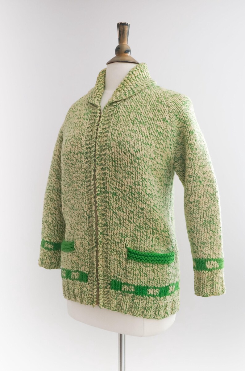 May include: A green and cream-colored knitted cardigan with a collared neckline and a full zipper. The sweater features green accents on the pockets and cuffs. The cardigan is displayed on a mannequin.