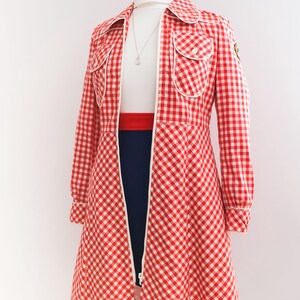 May include: A red and white gingham dress with a white collar and long sleeves. The dress has a zipper down the front and two pockets on the chest. The dress is on a mannequin.