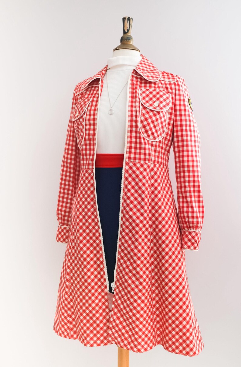 May include: A red and white gingham dress with a white collar and long sleeves. The dress has a zipper down the front and two pockets on the chest. The dress is on a mannequin.