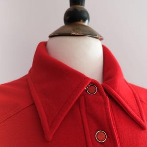 Op de afbeelding: Close-up van een felrood overhemd met kraag en twee ronde knopen. Het overhemd wordt op een mannequin gepresenteerd. De kraag is puntig en de knopen zijn zilverkleurig met rode centra. De stof lijkt van een gemiddeld gewicht te zijn.
