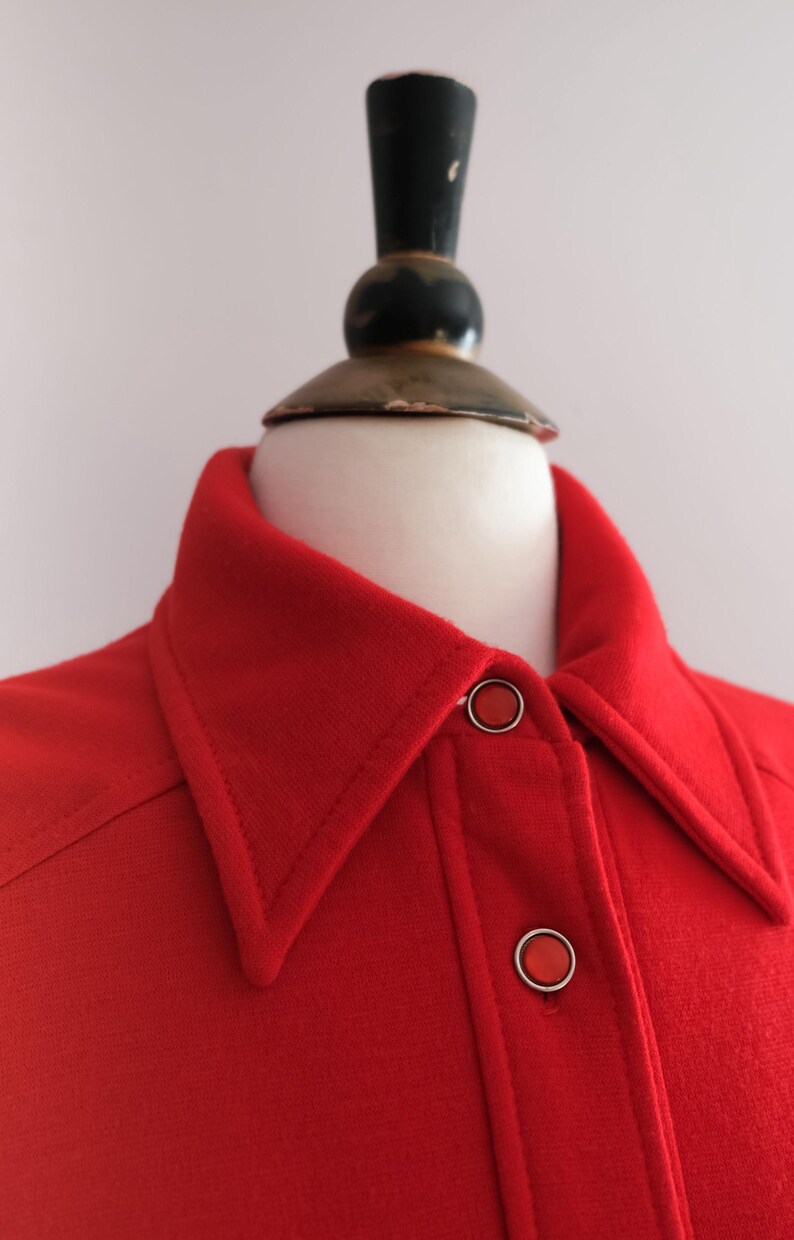 Op de afbeelding: Close-up van een felrood overhemd met kraag en twee ronde knopen. Het overhemd wordt op een mannequin gepresenteerd. De kraag is puntig en de knopen zijn zilverkleurig met rode centra. De stof lijkt van een gemiddeld gewicht te zijn.