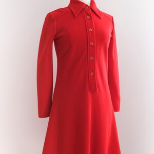 Op de afbeelding: Een felrode jurk met lange mouwen en een kraag. De jurk heeft een knoopsluiting aan de voorkant en een uitlopende rok, gepresenteerd op een mannequin. De jurk is effen van kleur, met een vintage stijl.