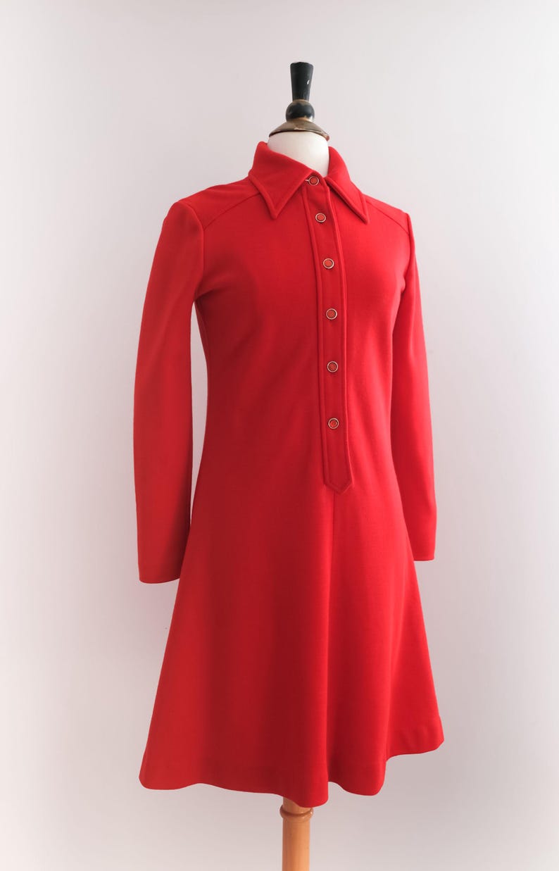 Op de afbeelding: Een felrode jurk met lange mouwen en een kraag. De jurk heeft een knoopsluiting aan de voorkant en een uitlopende rok, gepresenteerd op een mannequin. De jurk is effen van kleur, met een vintage stijl.