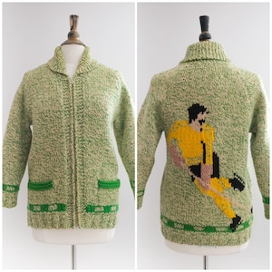 May include: A vintage green and beige knit cardigan with a zipper closure, two front pockets, and a green trim. The back features a yellow, black, and pink soccer player design. The sweater is displayed on a mannequin.