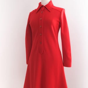 Op de afbeelding: Een felrode jurk met lange mouwen en een kraag. De jurk heeft een knoopsluiting aan de voorkant en een uitlopende rok, gepresenteerd op een mannequin. De jurk is effen van kleur en toont een klassieke, vintage stijl.
