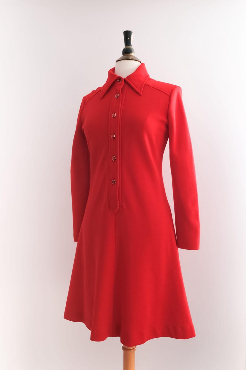 Op de afbeelding: Een felrode jurk met lange mouwen en een kraag. De jurk heeft een knoopsluiting aan de voorkant en een uitlopende rok, gepresenteerd op een mannequin. De jurk is effen van kleur en toont een klassieke, vintage stijl.