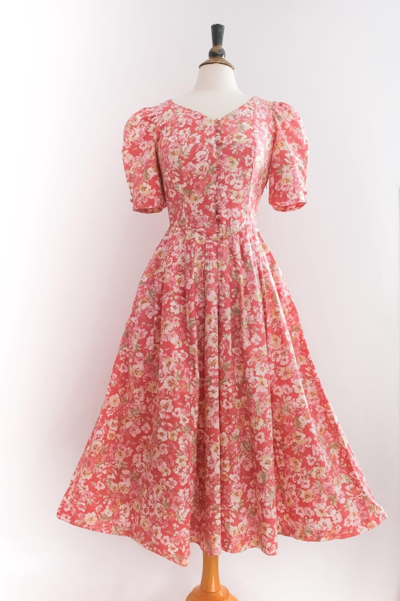 May include: A vintage-style, floral-print dress in shades of red, pink, and white. The dress features a V-neckline, short puffed sleeves, and a button-down bodice. The skirt is a full, flowing A-line, perfect for a retro look.