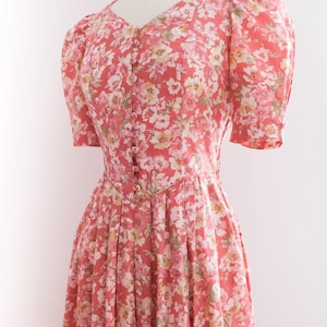 May include: A floral print dress in shades of coral, white, and yellow. The dress features a V-neckline, short puffed sleeves, and a button-down front. The skirt is pleated, creating a flowing silhouette. The dress is displayed on a mannequin.
