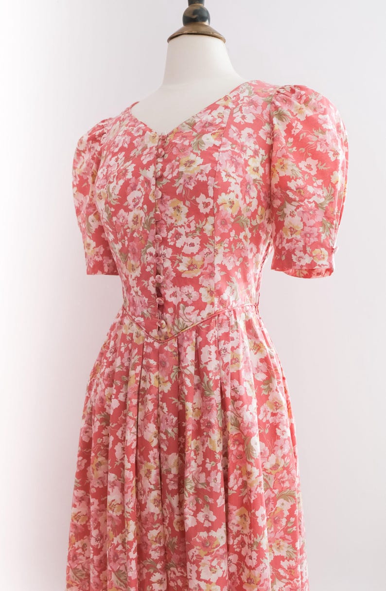May include: A floral print dress in shades of coral, white, and yellow. The dress features a V-neckline, short puffed sleeves, and a button-down front. The skirt is pleated, creating a flowing silhouette. The dress is displayed on a mannequin.