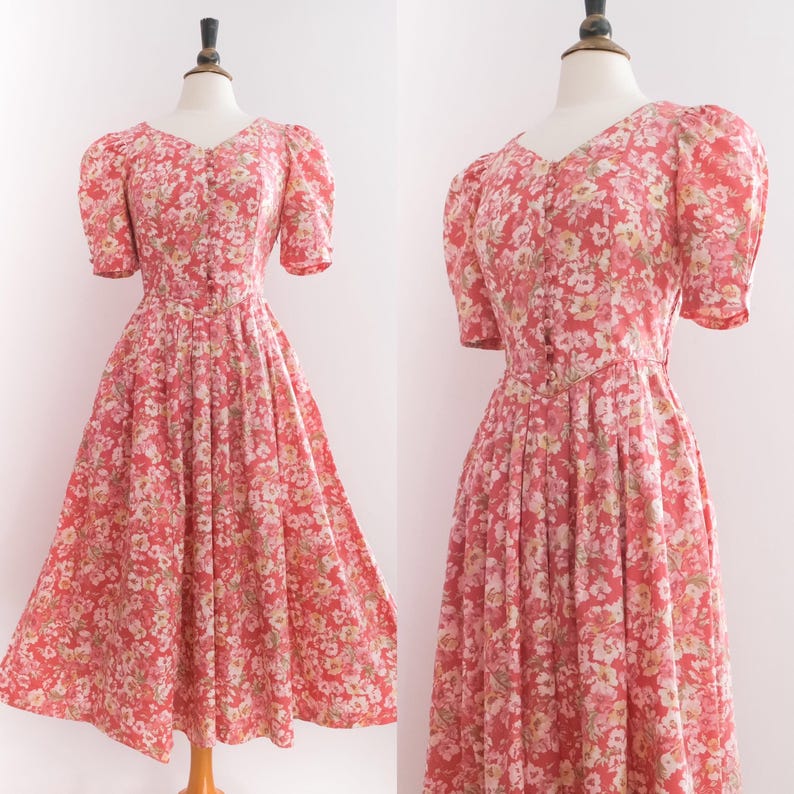 May include: A vintage-style, floral-print dress in shades of red, pink, and white. The dress features a V-neckline, short puffed sleeves, and a button-down bodice. The skirt is pleated and flows to a mid-calf length, creating a classic silhouette.