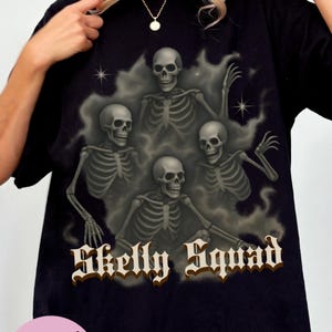 May include: Black t-shirt featuring a graphic of five skeletons in a smoky background with the text "Skelly Squad" in a gothic font. The shirt also has a pink circle with the text "Comfort Colors! (Black XL)".
