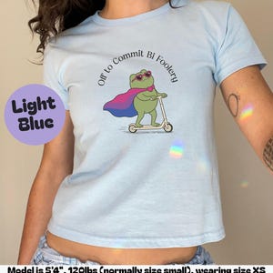 May include: A light blue t-shirt featuring a graphic of a green frog wearing pink heart-shaped sunglasses and a pink and purple cape while riding a white scooter. The text above the frog reads, Off to Commit Bi Foolery in black font.