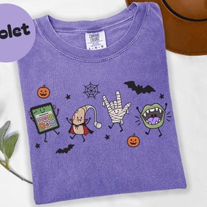 Könnte beinhalten: Ein violettes Kurzarm-T-Shirt mit einem Grafikdruck eines grünen Tablets, einer Hörgerätefigur mit Umhang, einer Mumienhand, die ein Zeichen macht, und einem grünen Mund mit Fangzähnen, umgeben von kleinen Kürbissen, Fledermäusen und einem Spinnennetz.
