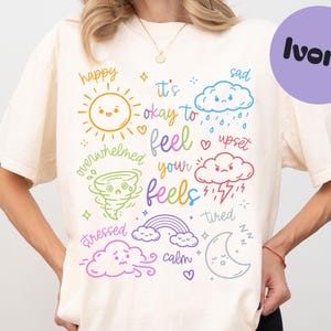 May include: An ivory t-shirt featuring a colorful graphic print with the text "it's okay to feel your feels" surrounded by illustrations of a sun, clouds, a tornado, a rainbow, and a moon, each labeled with various emotional states.